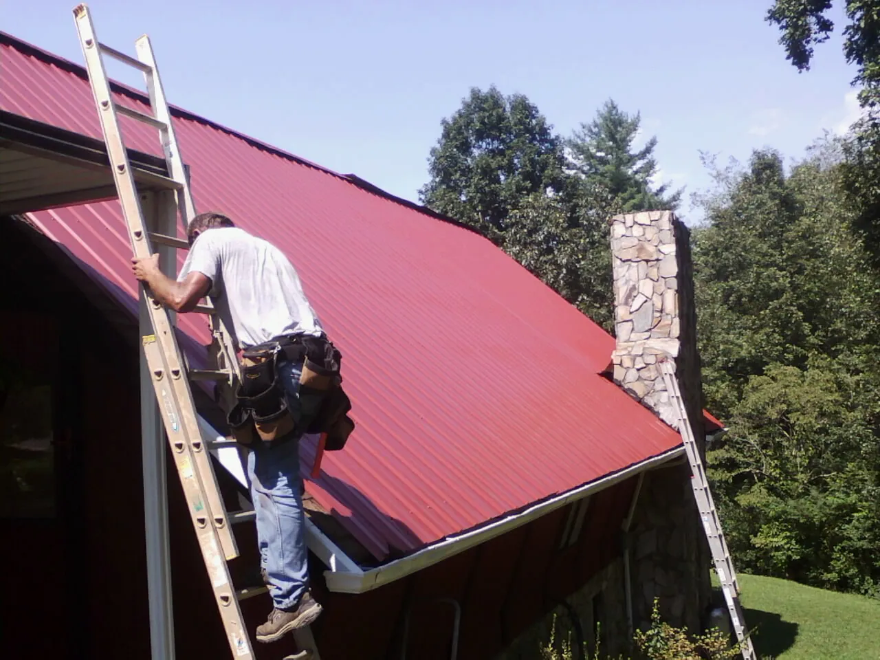 Roofer on a ladder beside a red metal roof and stone chimney — Soffit & Fascia Repair by Roofing Brothers in Boyd
